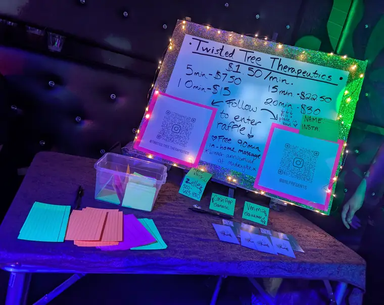 A table at a night event under string lights, set with a hand-lettered price board, a raffle box, printed cards and two QR codes